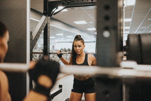 A woman is performing squats in a gym while facing a mirror. She is using a barbell with weights, and appears focused and determined. The gym environment is visible in the background with fitness equipment and fluorescent lighting.