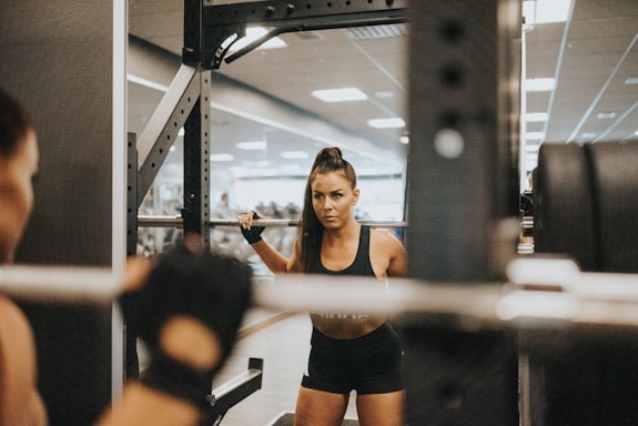 A woman is performing squats in a gym while facing a mirror. She is using a barbell with weights, and appears focused and determined. The gym environment is visible in the background with fitness equipment and fluorescent lighting.