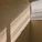 Sunlight filters through horizontal blinds, casting striped shadows along a beige wall. The window is partially open, revealing soft, diffused light and subtle reflections.