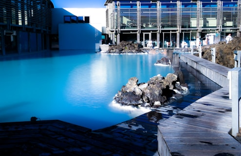 A serene, turquoise geothermal pool surrounded by black volcanic rocks and a modern building with large windows. A wooden walkway curves around the pool, while several people can be seen enjoying the thermal spa environment.