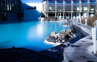 A serene, turquoise geothermal pool surrounded by black volcanic rocks and a modern building with large windows. A wooden walkway curves around the pool, while several people can be seen enjoying the thermal spa environment.