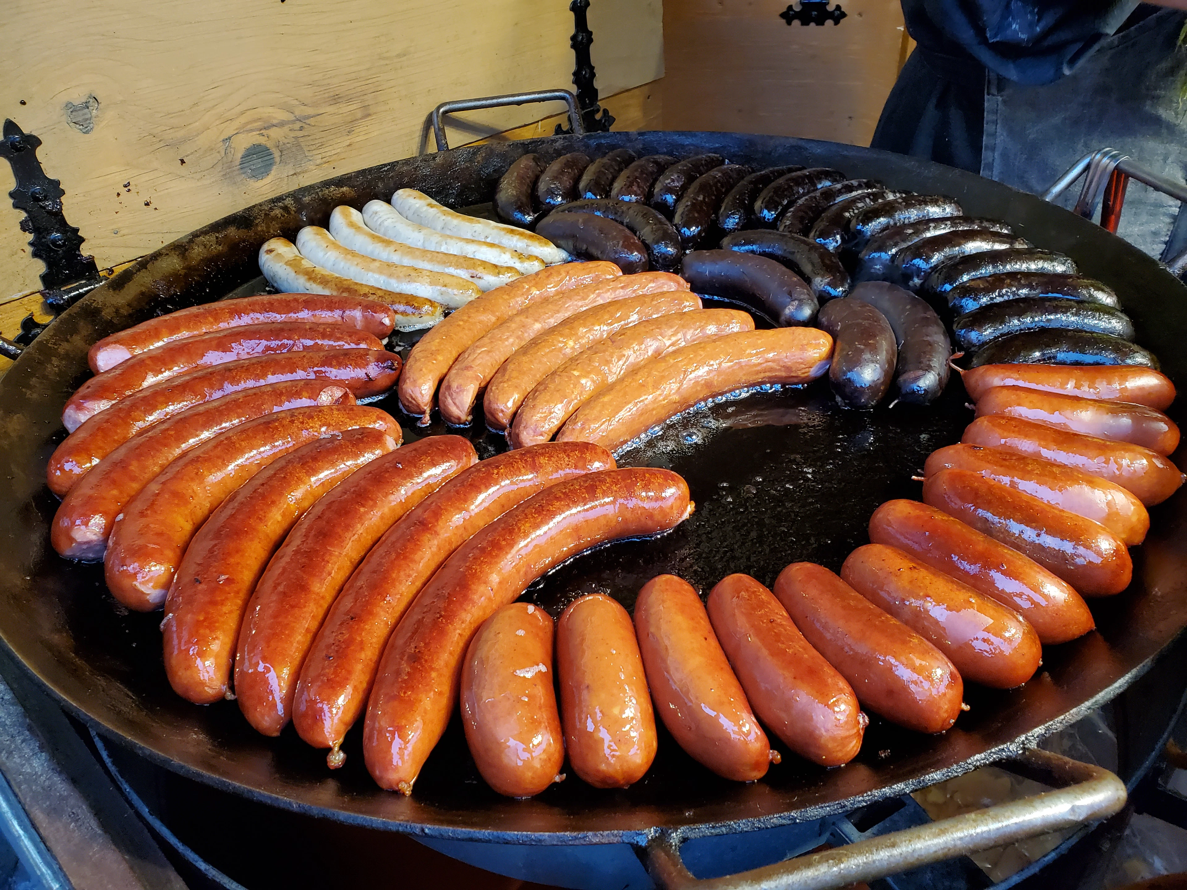 A wide iron griddle pan crowded with grilled sausages — pale white sausages, dark blood sausages and rings of smoked pork — sizzling at a Christmas market food stall in Tallinn, Estonia