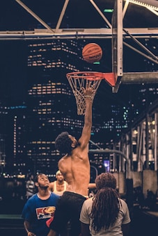 A group of people are playing basketball on an outdoor court at night. One person is shirtless, jumping to make a layup. The background features a cityscape with illuminated skyscrapers.