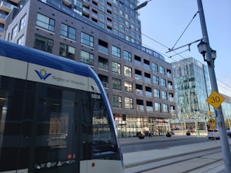 A street-level view features a light rail vehicle with the Region of Waterloo logo prominently displayed. The background includes a multi-story brick building with numerous windows and a more modern glass building. Street signs, a speed limit sign indicating 30, and overhead cables for the rail system are visible. The scene captures elements of urban transportation and architecture.