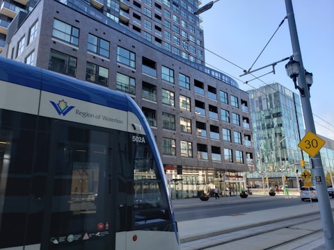 A street-level view features a light rail vehicle with the Region of Waterloo logo prominently displayed. The background includes a multi-story brick building with numerous windows and a more modern glass building. Street signs, a speed limit sign indicating 30, and overhead cables for the rail system are visible. The scene captures elements of urban transportation and architecture.