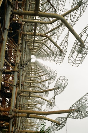 A large, rusted metal structure featuring a series of repeating, lattice-like elements that extend into the distance. The industrial framework is composed of thick, cylindrical supports that are interconnected by intricate metal mesh patterns.