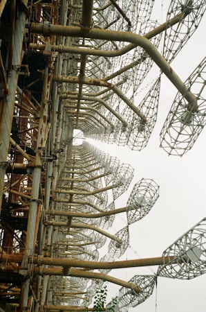 A large, rusted metal structure featuring a series of repeating, lattice-like elements that extend into the distance. The industrial framework is composed of thick, cylindrical supports that are interconnected by intricate metal mesh patterns.