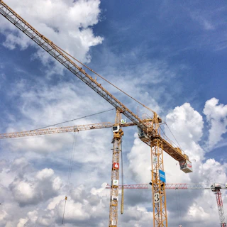 A towering crane lifting heavy steel beams against a deep blue sky at a bustling construction site.