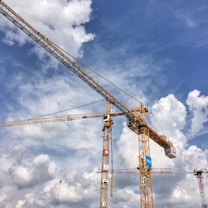 Tall construction cranes against a backdrop of a blue sky filled with scattered clouds. The cranes are yellow and positioned in a busy construction site environment.