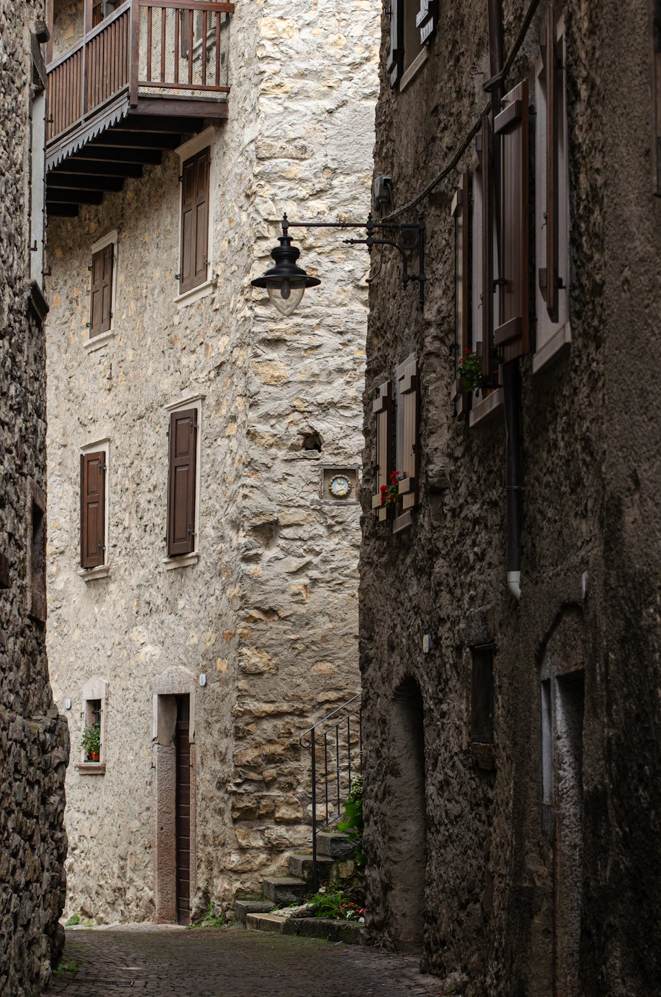 The image depicts a narrow cobblestone street flanked by old stone buildings with wooden shutters and balconies on both sides, creating an atmosphere of quaint charm. The perspective is from ground level, looking down the street towards the horizon line, giving a sense of depth to the scene. A solitary black lamppost stands in the foreground, adding a touch of modernity to the otherwise traditional setting.