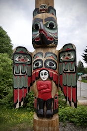 A totem pole with intricate carvings stands amidst green foliage. The pole features a large bird with outstretched wings, decorated with bold red, black, and green patterns. Below the bird's beak, there is a human figure with pale skin, wearing a red shirt and black pants, arms crossed. Above the bird's head and on its wings are smaller human faces with distinct expressions.