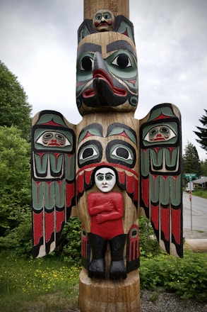 A totem pole with intricate carvings stands amidst green foliage. The pole features a large bird with outstretched wings, decorated with bold red, black, and green patterns. Below the bird's beak, there is a human figure with pale skin, wearing a red shirt and black pants, arms crossed. Above the bird's head and on its wings are smaller human faces with distinct expressions.