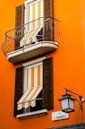 An orange building facade with two windows featuring striped orange and white awnings. Both windows have wooden shutters, and the upper window includes a small balcony with black metal railing. A street sign labeled 'Via V. Emanuele' is mounted on the wall beside an ornate black street lamp.