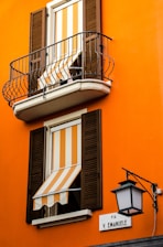 An orange building facade with two windows featuring striped orange and white awnings. Both windows have wooden shutters, and the upper window includes a small balcony with black metal railing. A street sign labeled 'Via V. Emanuele' is mounted on the wall beside an ornate black street lamp.