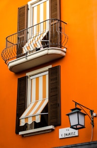 An orange building facade with two windows featuring striped orange and white awnings. Both windows have wooden shutters, and the upper window includes a small balcony with black metal railing. A street sign labeled 'Via V. Emanuele' is mounted on the wall beside an ornate black street lamp.