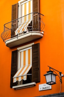 An orange building facade with two windows featuring striped orange and white awnings. Both windows have wooden shutters, and the upper window includes a small balcony with black metal railing. A street sign labeled 'Via V. Emanuele' is mounted on the wall beside an ornate black street lamp.
