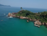 A small, lush, green island surrounded by clear, turquoise water. The island features rocky formations and a few tall palm trees. A wooden pier extends into the water with small boats nearby. In the background, hazy mountains can be seen on the horizon under a clear blue sky.