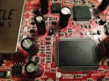 A close-up view of a red printed circuit board featuring numerous integrated circuits, capacitors, and resistors. The board is populated with various electronic components, including black cylindrical capacitors and rectangular black chips with many pins. The text 'PINNACLE SYSTEMS' is visible, along with various alphanumeric identifiers.