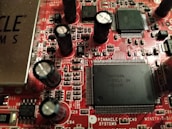 A close-up view of a red printed circuit board featuring numerous integrated circuits, capacitors, and resistors. The board is populated with various electronic components, including black cylindrical capacitors and rectangular black chips with many pins. The text 'PINNACLE SYSTEMS' is visible, along with various alphanumeric identifiers.