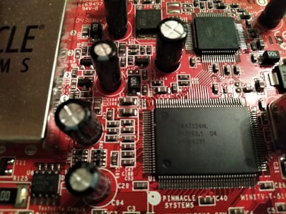 A close-up view of a red printed circuit board featuring numerous integrated circuits, capacitors, and resistors. The board is populated with various electronic components, including black cylindrical capacitors and rectangular black chips with many pins. The text 'PINNACLE SYSTEMS' is visible, along with various alphanumeric identifiers.