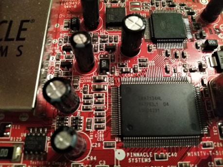 A close-up view of a red printed circuit board featuring numerous integrated circuits, capacitors, and resistors. The board is populated with various electronic components, including black cylindrical capacitors and rectangular black chips with many pins. The text 'PINNACLE SYSTEMS' is visible, along with various alphanumeric identifiers.