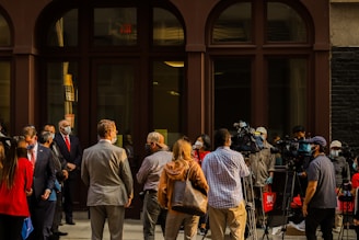 A group of people, including several wearing masks, are gathered outside a building. Some are in business attire, suggesting a formal event or press conference. Several individuals with cameras and tripods are positioned towards the center, indicating media coverage.