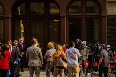 A group of people, including several wearing masks, are gathered outside a building. Some are in business attire, suggesting a formal event or press conference. Several individuals with cameras and tripods are positioned towards the center, indicating media coverage.