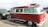 A vintage red and cream Volkswagen van is parked in a lot, featuring a retro design with a checkered stripe along the base. The windows have decorative curtains, and a roof rack holds a metal storage unit. Nearby are other cars, including a dark SUV and another vehicle.