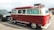 A vintage red and cream Volkswagen van is parked in a lot, featuring a retro design with a checkered stripe along the base. The windows have decorative curtains, and a roof rack holds a metal storage unit. Nearby are other cars, including a dark SUV and another vehicle.