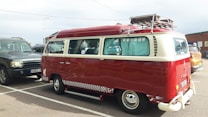 A vintage red and cream Volkswagen van is parked in a lot, featuring a retro design with a checkered stripe along the base. The windows have decorative curtains, and a roof rack holds a metal storage unit. Nearby are other cars, including a dark SUV and another vehicle.