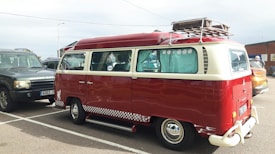 A vintage red and cream Volkswagen van is parked in a lot, featuring a retro design with a checkered stripe along the base. The windows have decorative curtains, and a roof rack holds a metal storage unit. Nearby are other cars, including a dark SUV and another vehicle.