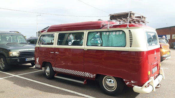A vintage red and cream Volkswagen van is parked in a lot, featuring a retro design with a checkered stripe along the base. The windows have decorative curtains, and a roof rack holds a metal storage unit. Nearby are other cars, including a dark SUV and another vehicle.
