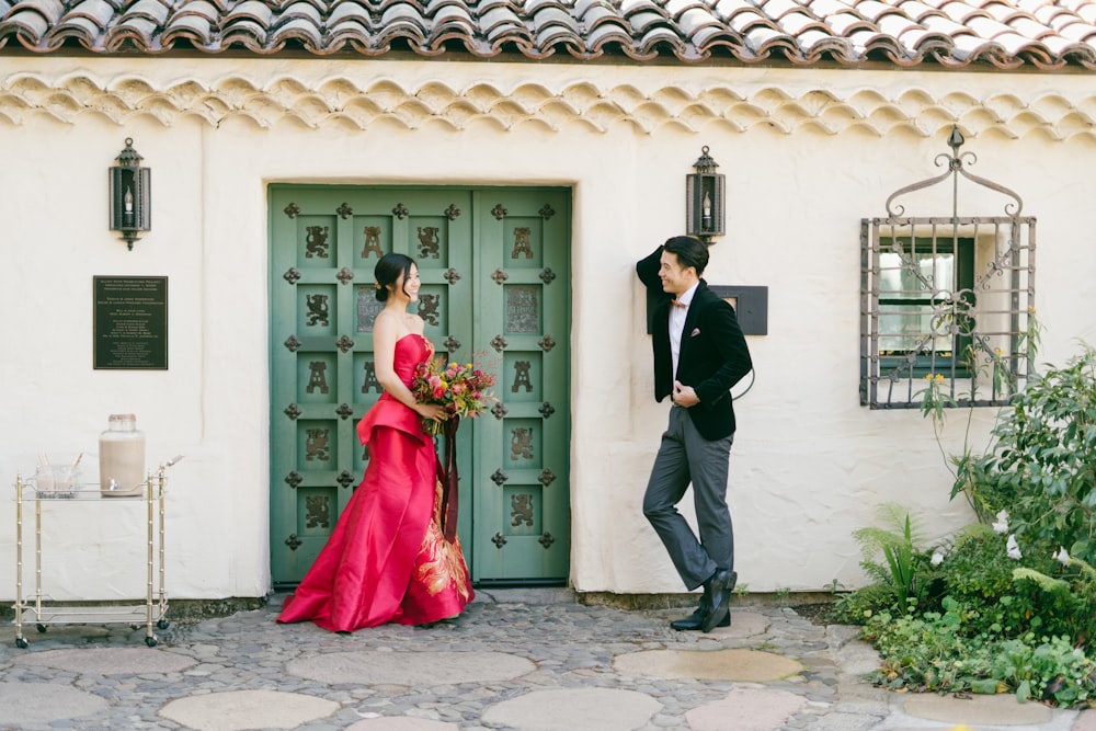 homem na jaqueta do terno preto e mulher no vestido vermelho em pé perto da porta de madeira branca durante