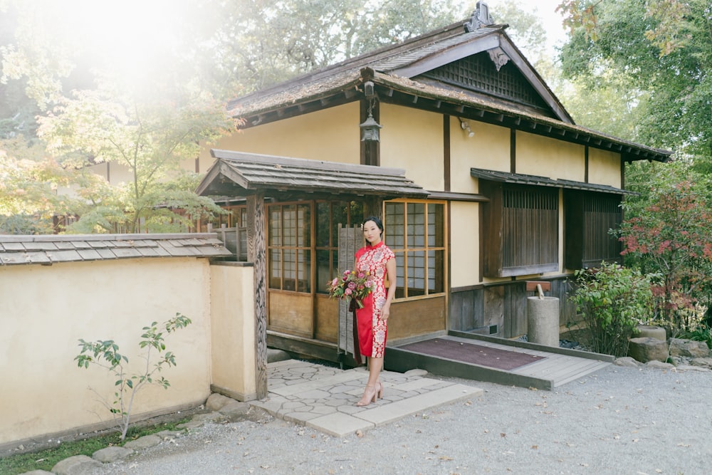赤と白の花柄のドレスを着た女性が、昼間、茶色の木造家屋のそばに立つ