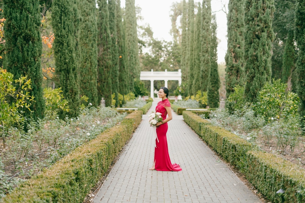 woman in red dress walking on pathway between green plants during daytime