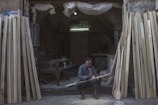 A man sits on the ground at the entrance of a workshop, focused on carving a piece of wood. The workshop is filled with various wooden planks leaning against the walls. The environment is dimly lit, with a single bulb providing light inside the space. The man is surrounded by sawdust and uses a tool to shape the wood. He is wearing a plaid shirt and appears to be deeply engrossed in his work.