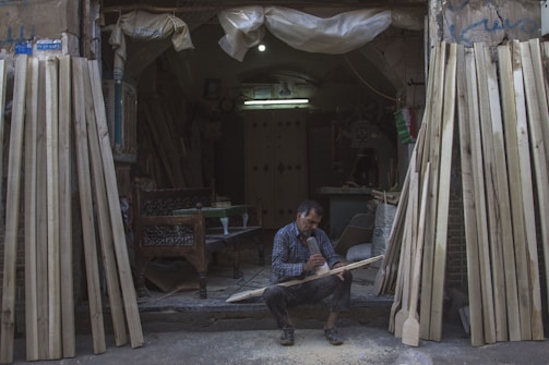 A man sits on the ground at the entrance of a workshop, focused on carving a piece of wood. The workshop is filled with various wooden planks leaning against the walls. The environment is dimly lit, with a single bulb providing light inside the space. The man is surrounded by sawdust and uses a tool to shape the wood. He is wearing a plaid shirt and appears to be deeply engrossed in his work.