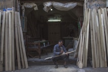 A man sits on the ground at the entrance of a workshop, focused on carving a piece of wood. The workshop is filled with various wooden planks leaning against the walls. The environment is dimly lit, with a single bulb providing light inside the space. The man is surrounded by sawdust and uses a tool to shape the wood. He is wearing a plaid shirt and appears to be deeply engrossed in his work.