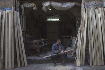 A man sits on the ground at the entrance of a workshop, focused on carving a piece of wood. The workshop is filled with various wooden planks leaning against the walls. The environment is dimly lit, with a single bulb providing light inside the space. The man is surrounded by sawdust and uses a tool to shape the wood. He is wearing a plaid shirt and appears to be deeply engrossed in his work.