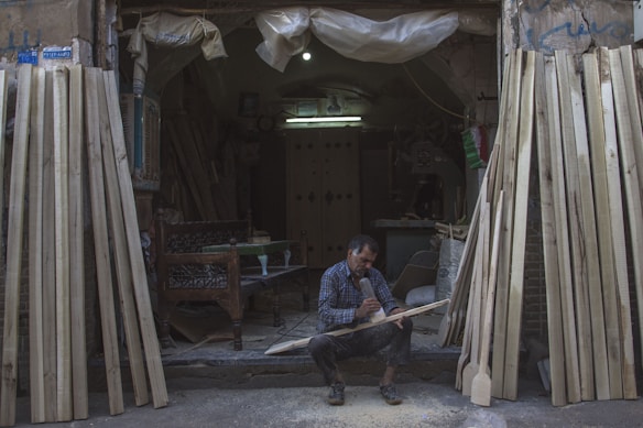 A man sits on the ground at the entrance of a workshop, focused on carving a piece of wood. The workshop is filled with various wooden planks leaning against the walls. The environment is dimly lit, with a single bulb providing light inside the space. The man is surrounded by sawdust and uses a tool to shape the wood. He is wearing a plaid shirt and appears to be deeply engrossed in his work.