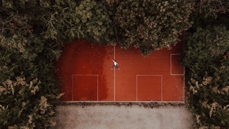 Aerial view of a red sports court surrounded by dense green trees, with a person lying in the center of the court. The court has rectangular markings, indicating it might be used for multiple sports.
