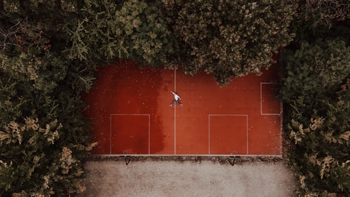 Aerial view of a red sports court surrounded by dense green trees, with a person lying in the center of the court. The court has rectangular markings, indicating it might be used for multiple sports.