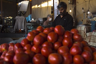 A market scene where a mound of ripe red tomatoes is prominently displayed in the foreground. In the background, two men can be seen; one is wearing a mask and operating a machine, while the other is focused on sorting or organizing the produce. The setting appears to be a dimly lit indoor market with various crates and bulbs hanging from above.
