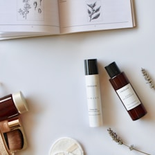 A flat lay composition features an open book with botanical illustrations, several skincare products in elegant bottles, a makeup brush, cotton pads, and sprigs of lavender arranged on a minimalist white background.