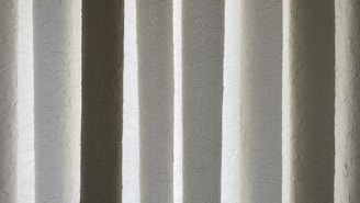 Vertical blinds with a crinkled texture appear as alternating light and dark bands. The material has a subtle sheen and displays a mix of soft white, gray, and beige colors.