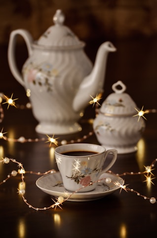 A beautifully crafted set of porcelain teaware consisting of a teapot, a sugar bowl, and a teacup with a saucer. The set features delicate floral patterns. Warm fairy lights are artistically arranged around the teaware, adding a cozy and enchanting atmosphere.