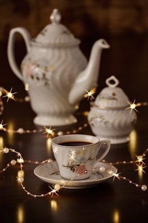 A beautifully crafted set of porcelain teaware consisting of a teapot, a sugar bowl, and a teacup with a saucer. The set features delicate floral patterns. Warm fairy lights are artistically arranged around the teaware, adding a cozy and enchanting atmosphere.