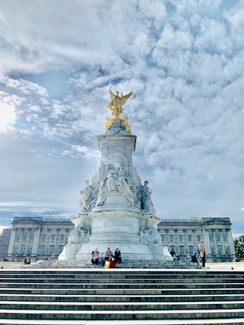 A large stone monument featuring intricate sculptures and a gilded angel at the top. The background includes a large classical building with many windows, and broad steps leading up to the monument with people sitting on them. The sky is expansive and partly cloudy.