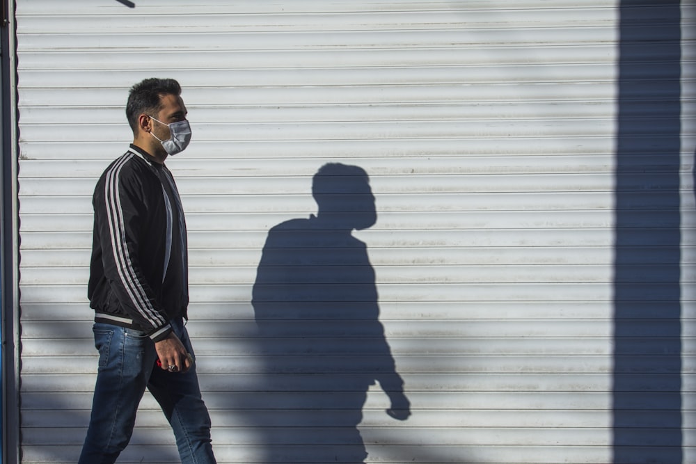 man in black jacket and blue denim jeans standing beside white wall