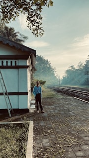 A person stands on a platform near a railway track, surrounded by lush greenery and trees. The building next to them has a ladder leaning against it, and the ground is paved with stones. The atmosphere is misty with a bluish tint, suggesting an early morning setting.